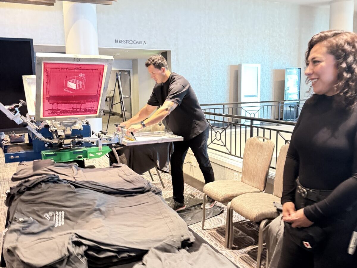 Merch Troop printer working the press while a guest laughs beside the shirt stack