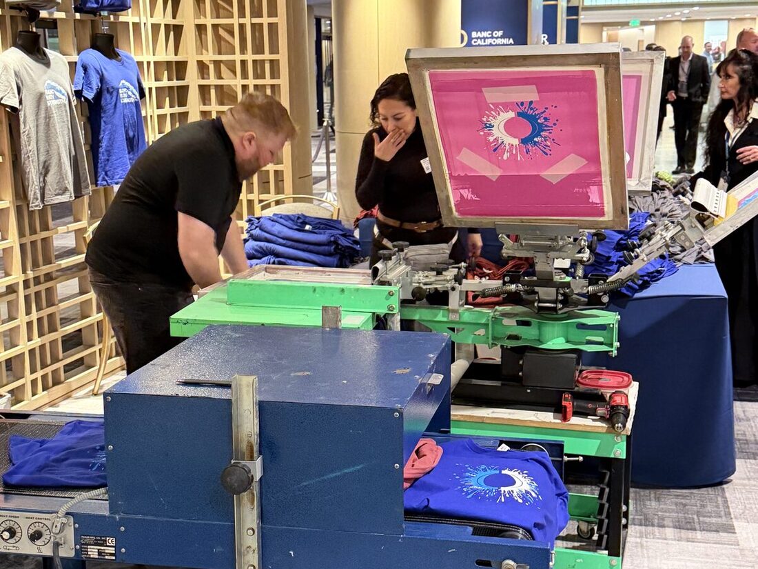 Bright pink screen loaded on the press with fresh shirts stacked at a live Merch Troop station